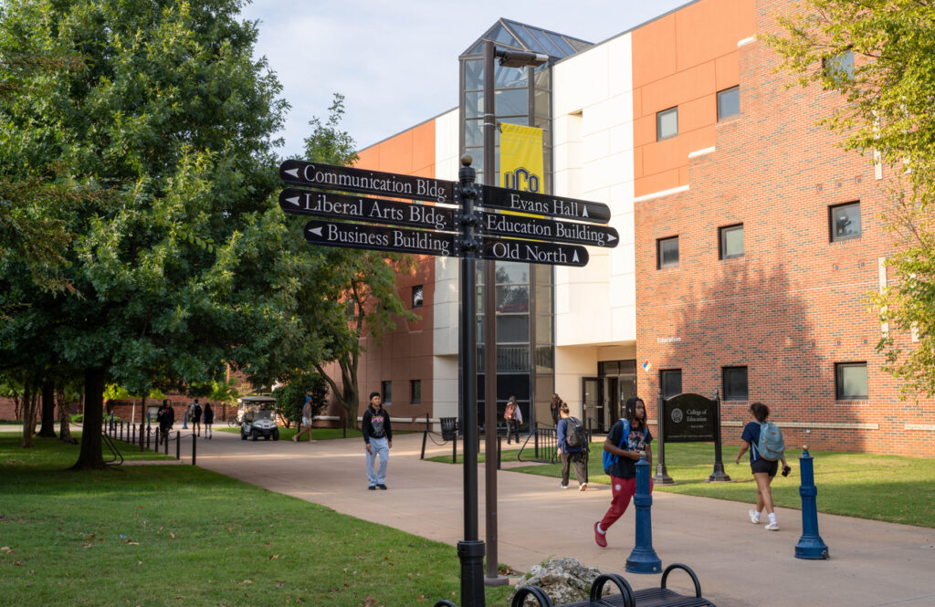 EDMOND, OKLAHOMA, USA - SEPTEMBER 8, 2025: A blue glass historical marker stands on the campus of the University of Central Oklahoma, featuring the university seal and a quote by President Don Betz. In the background, the iconic red-brick Old North building is visible through the summer foliage.