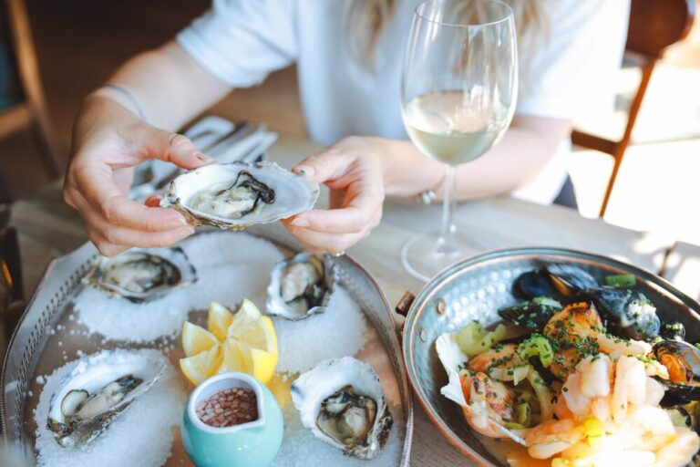 Hands of woman holding glass of champagne and fresh oyster with lemon juice in fish restaurant, selective focus. Seafood, French cuisine, fine dining concept