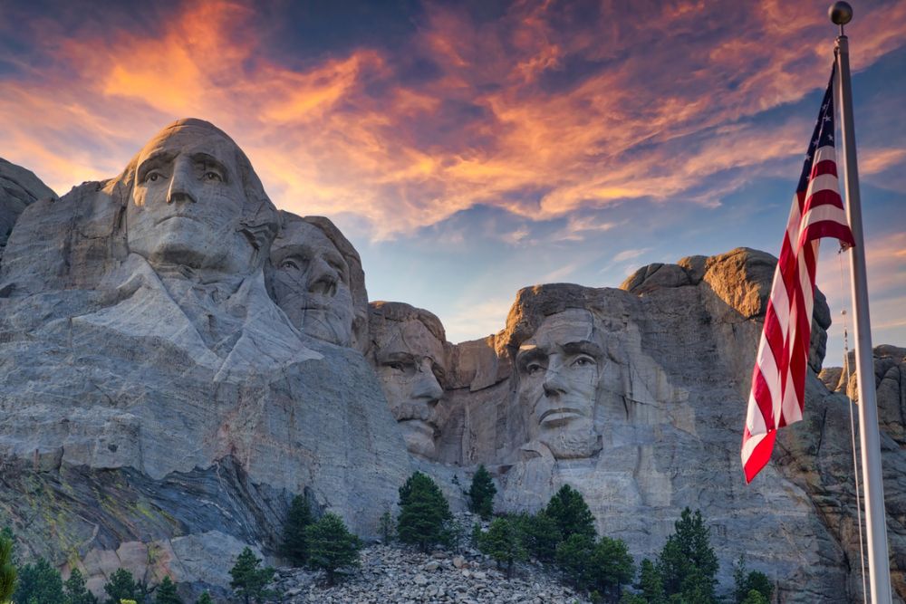 An American flag waves in front of Mount Rushmore National Memorial. The carved faces of four US presidents at the Mount Rushmore National Memorial illuminated at dusk.