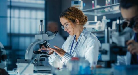 Hisplanic Female Bioengineer Looking at DNA Samples Under a Microscope in a Modern Applied Science Laboratory. Portrait of a Young Lab Engineer in White Coat Inventing New Medical Drugs