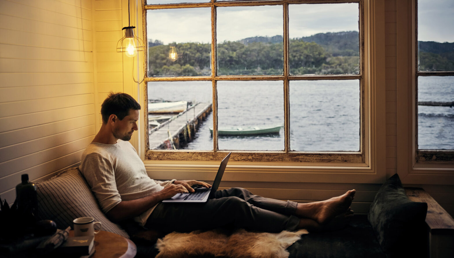 A man working on his laptop in front of a window overlooking a boat dock