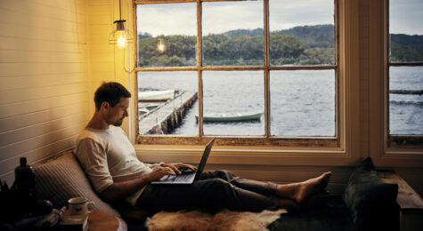 A man working on his laptop in front of a window overlooking a boat dock