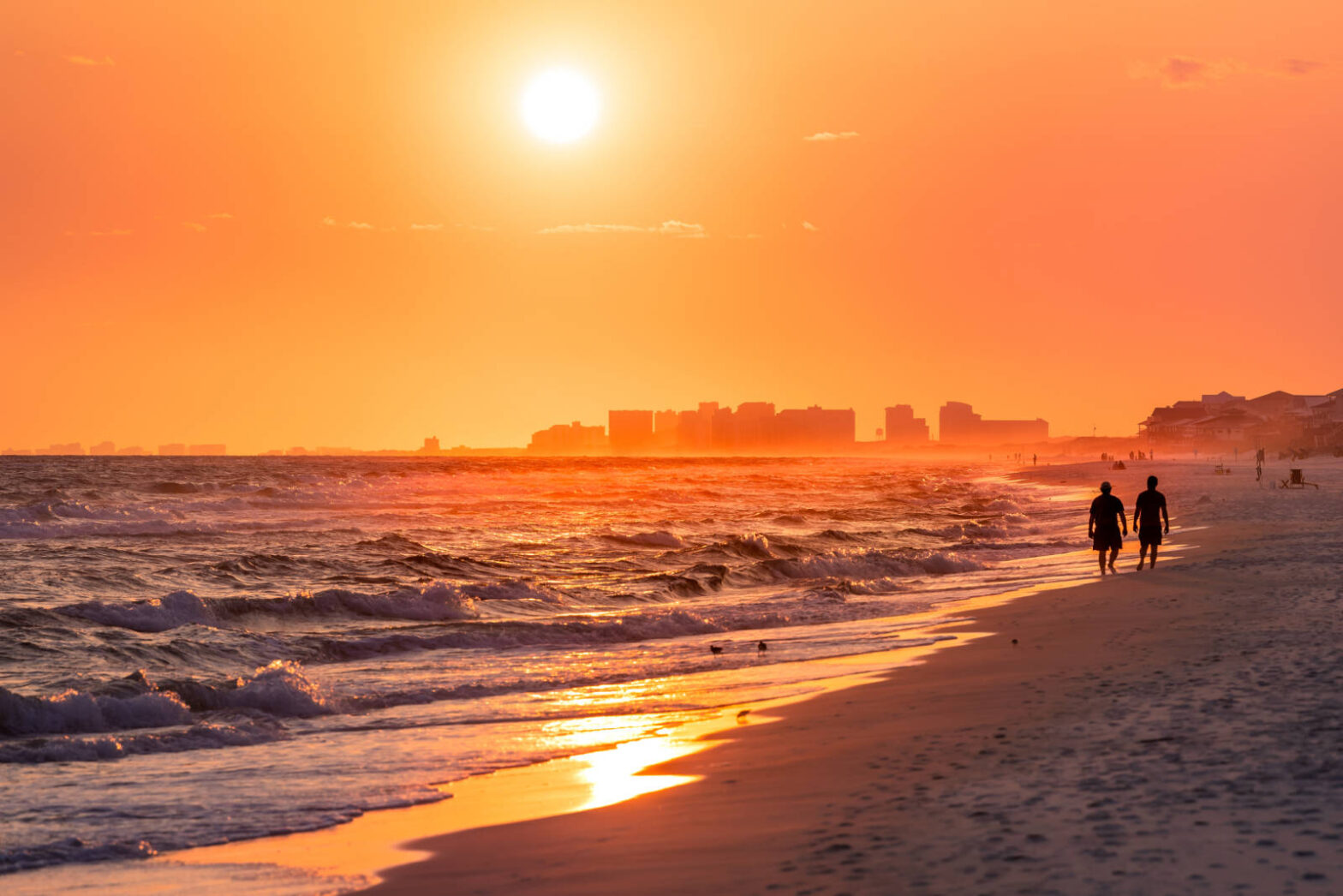 Couple walking on shore with dramatic orange red yellow sunset in Santa Rosa Beach, Florida with Pensacola coastline coast cityscape skyline in panhandle with ocean gulf mexico waves