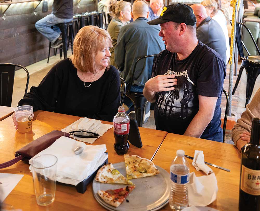 People eating pizza and drinking craft beer inside a modern brick-walled brewery taproom.