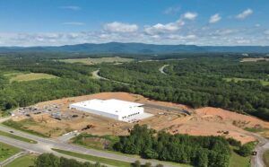 Aerial view of a large white roofed warehouse under construction surrounded by dirt and woods with mountains in background.