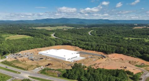 Aerial view of a large white roofed warehouse under construction surrounded by dirt and woods with mountains in background.