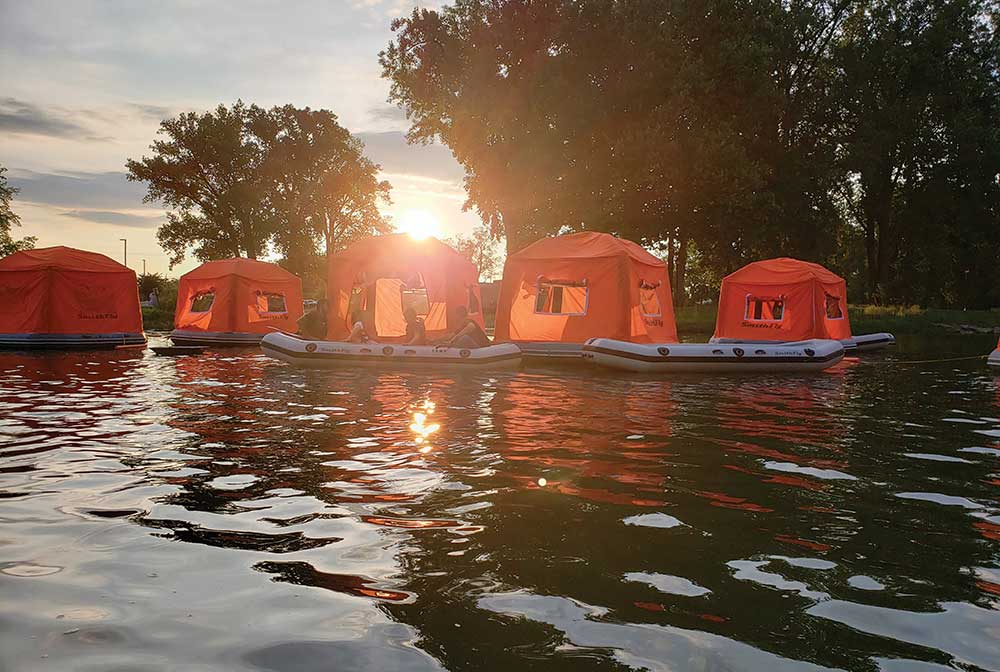 Brightly colored inflatable camping tents floating on calm river water near a grassy shoreline.