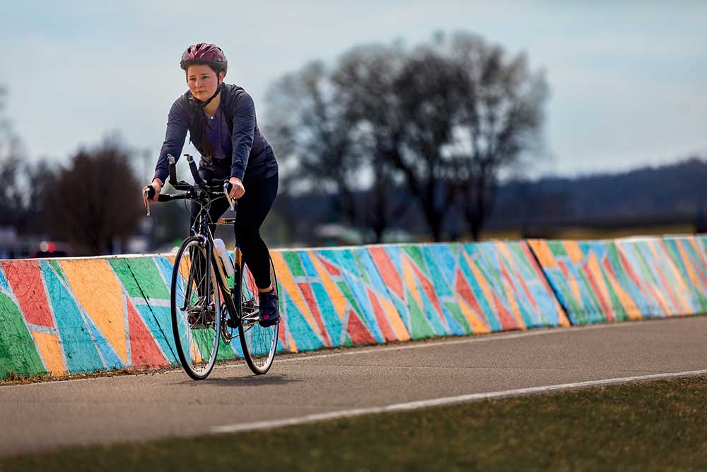 A person wearing a helmet riding a bicycle on a paved greenway trail through a park.