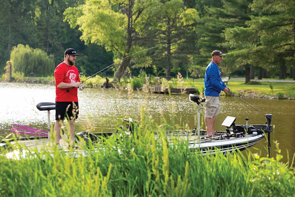 Two people fishing with fishing rods along a grassy lake bank on a sunny day.