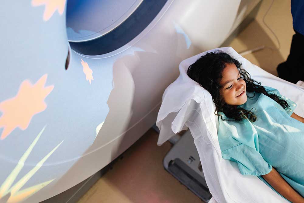 A smiling young girl in a light blue hospital gown lying on a white medical imaging table.
