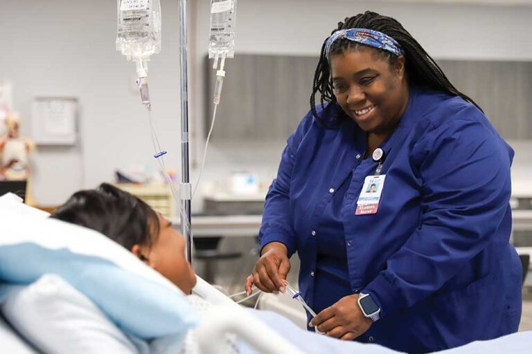 A student wearing scrubs practicing clinical procedures on a medical mannequin in a simulation lab.