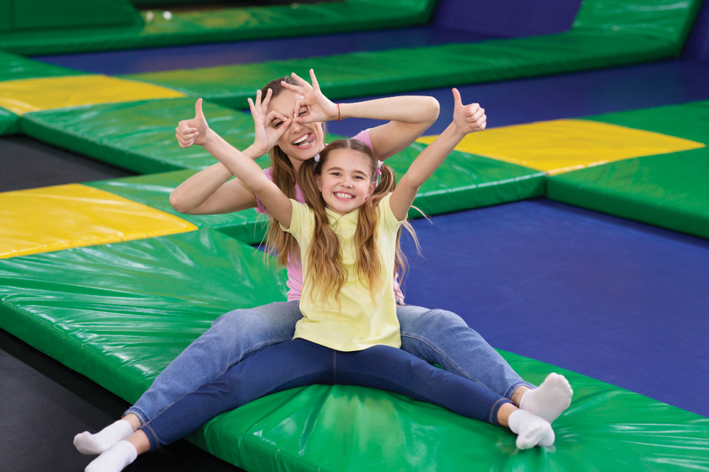 Playful silly mom with her teen daughter sitting at trampoline area of entertainment centre, showing thumbs up gesture. Mother and her child recommending to visit indoor kids playground