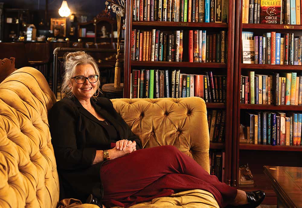 Danniele Reeves smiling on a gold tufted velvet chaise in front of tall wooden bookshelves at The Raven in Brentwood, Tennessee.