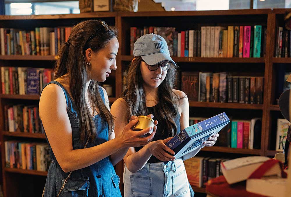 Two young women looking at a book and a candle beside a table of gift-wrapped books and mushroom decor at The Raven bookstore in Brentwood, Tennessee