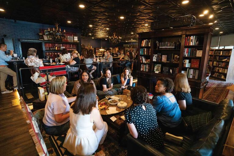 Groups of guests chatting at tables beside bookshelves and a dimly lit bar inside The Raven bookstore bar in Brentwood, TN
