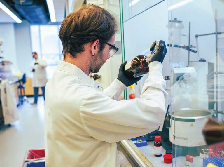 Scientist in a white lab coat and gloves examining a sample bottle in a research lab at the Warren Center for Neuroscience Drug Discovery in Franklin, Tennessee