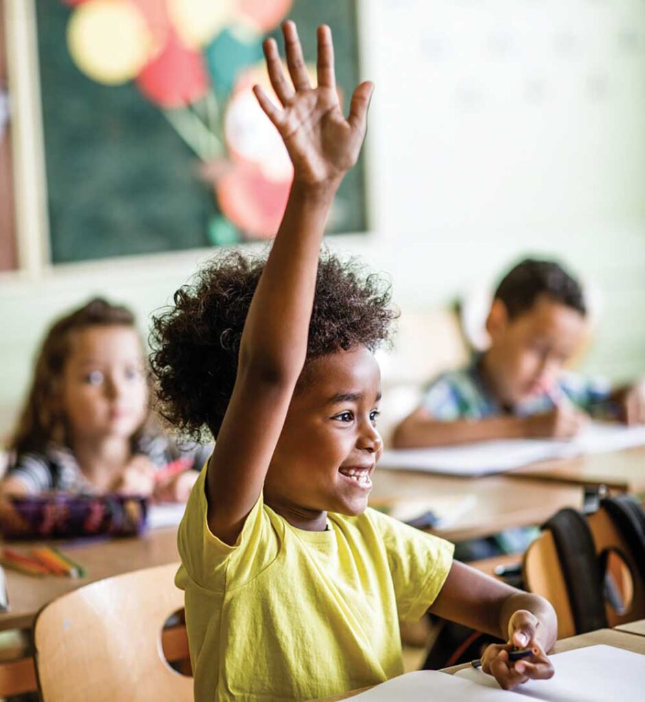 Smiling young child in a yellow shirt eagerly raising her hand at her desk in an elementary classroom.