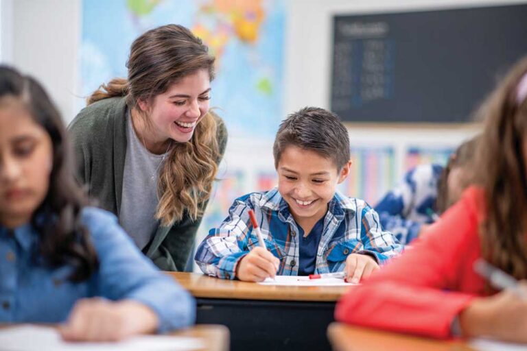 Smiling teacher leaning over to help an elementary student in a plaid shirt with his writing at a classroom table
