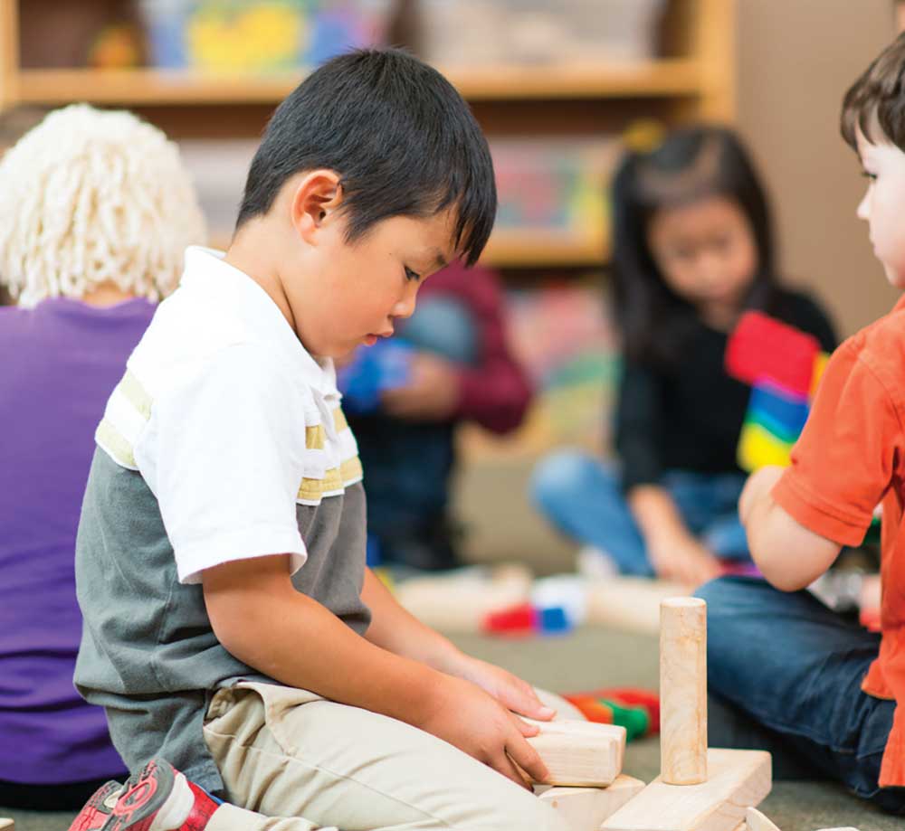 Young boy sitting on a classroom carpet stacking wooden building blocks, with other preschoolers playing in the background